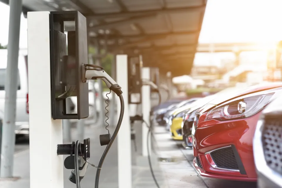 Close-up of red car parked and plugged in at EV charging station with row of cars behind it.